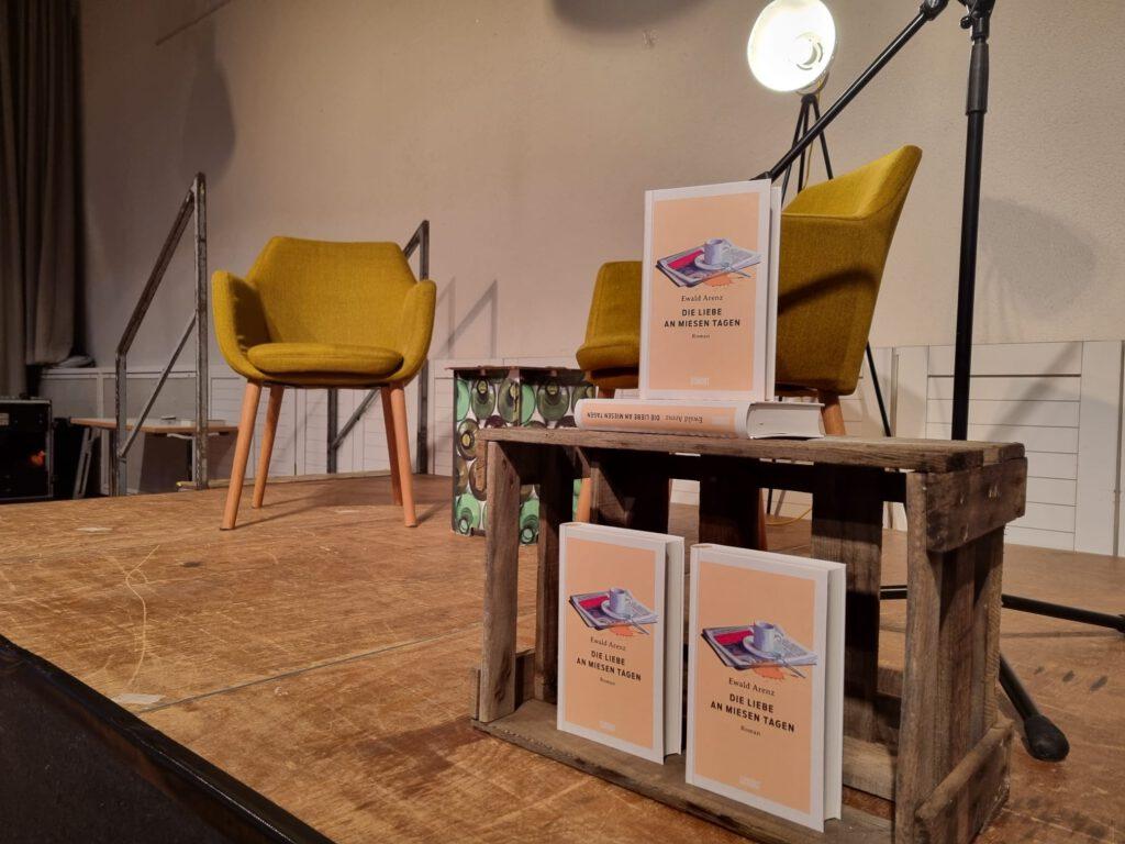 Foto von Bühne auf der gelbe Stühle, ein Mikro, eine Stehlampe und eine Holzkiste mit mehreren Ausgaben von Ewald Arenz Bestseller „Die Liebe an miesen Tagen“ drauf stehen.