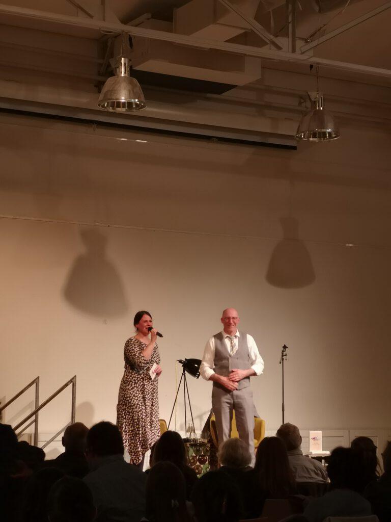 Foto von Ewald Arenz auf der Bühne und Elena Riedel seiner Lesung wie sie gerade zum Publikum sprechen.
