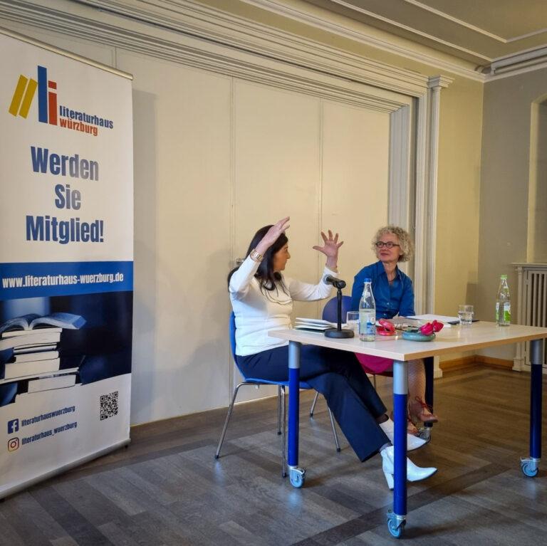 Ein Foto von Hannelore Schlaffer wie sie neben Regina Frisch vorne vor Publikum am Tisch sitzt und gestikuliert, links im Bild ein L-Banner vom Literaturhaus Würzburg mit der Aufschrift "Werden Sie Mitglied!"