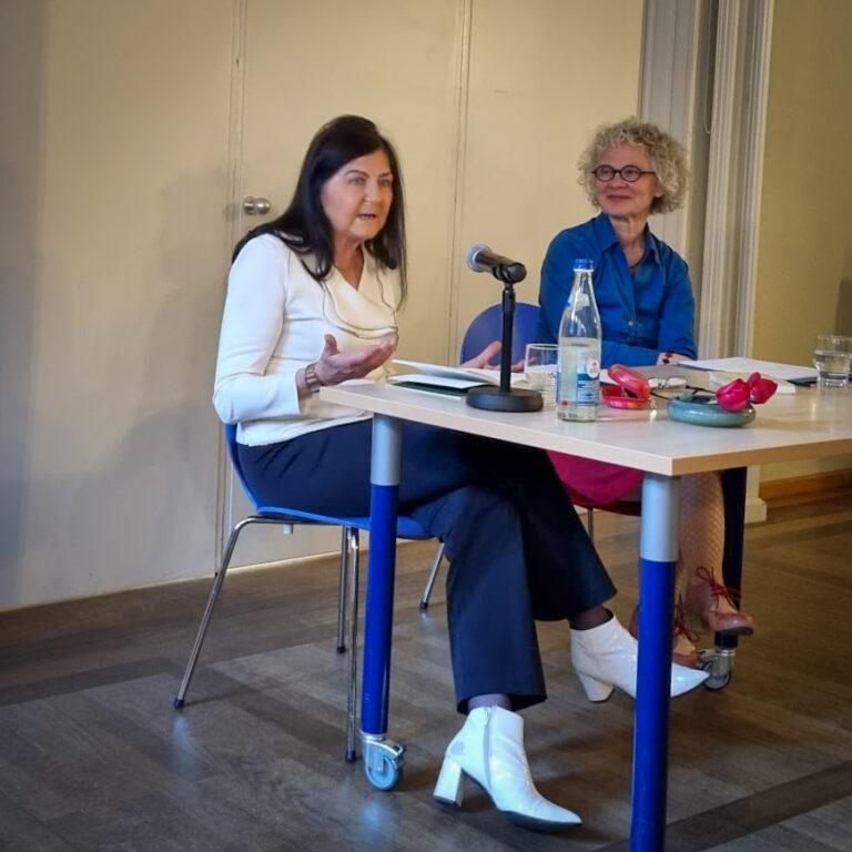 Ein Foto von Hannelore Schlaffer wie sie neben Regina Frisch vorne vor Publikum am Tisch sitzt und ins Mikro spricht.